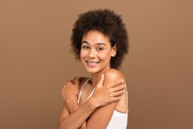 Photo cheerful millennial african american curly lady in white top with perfect skin and nude makeup hug her shoulders