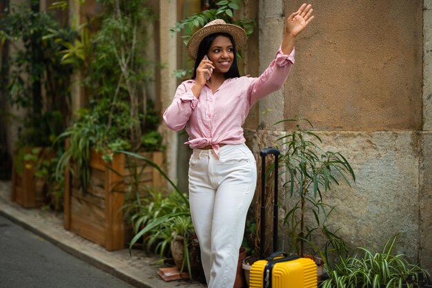 Photo cheerful millennial african american woman tourist in hat with suitcase calling by phone waving hand enjoy travel