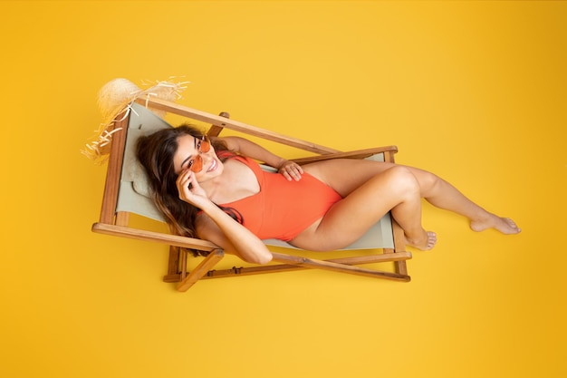 Cheerful millennial asian woman in swimsuit and sunglasses sit on deck chair enjoy free time