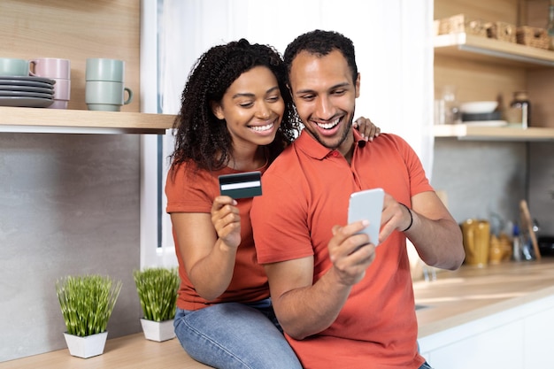 Cheerful millennial black lady hugging guy with credit card and smartphone enjoy online shopping in kitchen