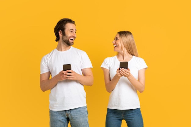 Cheerful millennial caucasian female and male in white tshirts look at each other hold phones