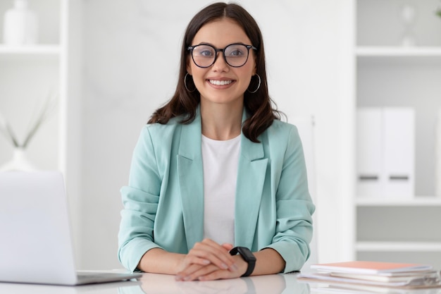 Cheerful millennial caucasian lady secretary manager in suit and glasses at workplace with laptop