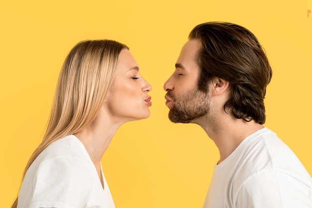 Cheerful millennial caucasian man and blonde lady in white tshirts are kissing isolated on yellow background