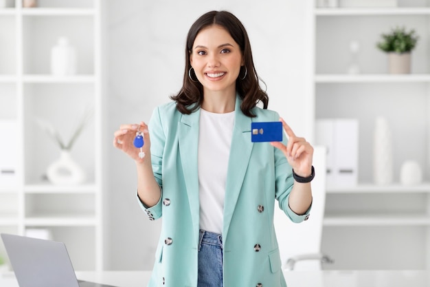 Cheerful millennial european businesswoman recommends credit card house keys in white office