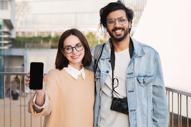 Веселый индийский парень-миллениал обнимает кавказскую леди, показывает смартфон с пустым экраном, рекомендует приложение