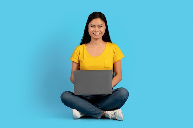 Cheerful millennial korean woman student blogger in yellow tshirt sit on floor with laptop watch video