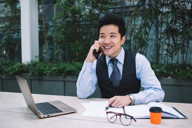 Cheerful modern Asian businessman speaking on phone on terrace