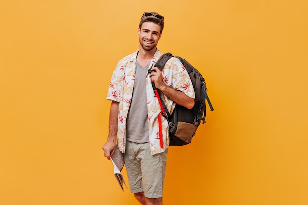 Cheerful modern man with ginger beard in grey tshirt and printed shirt smiling looking into camera and posing with large backpack