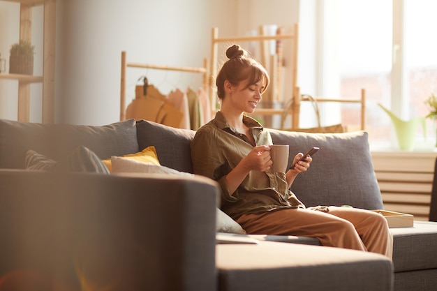 Cheerful modern young woman with hair bun sitting on sofa and reading internet news over coffee