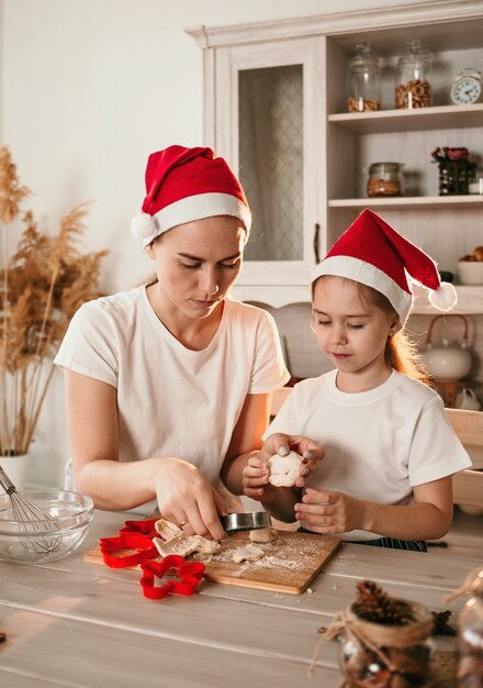 クリスマスの帽子をかぶった陽気なママと娘がキッチンのテーブルで遊ぶ