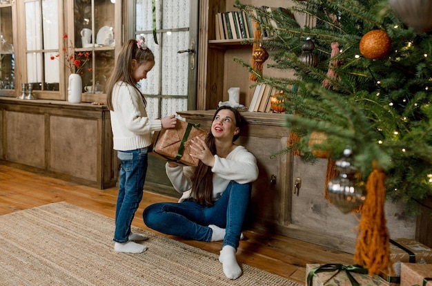 쾌활한 엄마와 그녀의 귀여운 딸이 선물을 교환합니다. 부모와 어린 아이가 실내 나무 근처에서 즐거운 시간을 보내고 있습니다. 방에 선물을 들고 사랑하는 가족