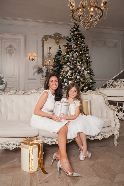 Cheerful mom and her cute daughter girl exchanging gifts in white classic interior against the background of a piano and a decorated Christmas tree.