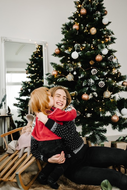 Cheerful mom kissing cute baby daughter girl near christmas\
tree merry christmas and happy holidays mother and little child\
having fun and playing together at home close up