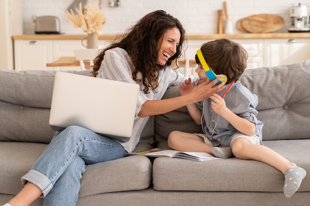 Cheerful mom and son laugh together on sofa mother work on laptop and kid listen to audio lesson