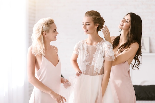 Cheerful moment. Delighted smiling young bridesmaids standing in the white bedroom while helping the bride to get ready and expressing happiness