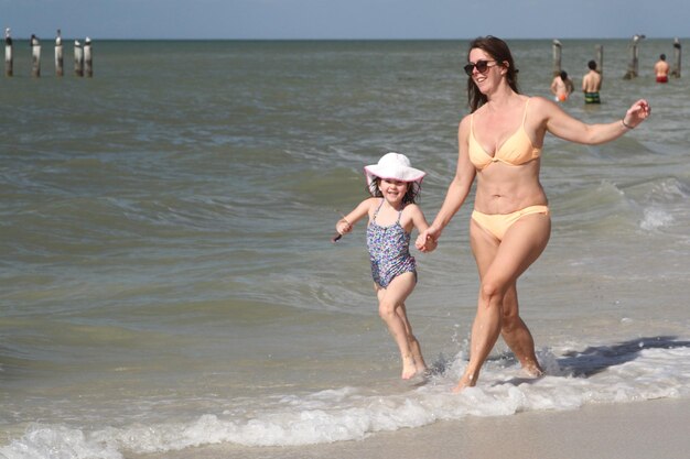 Photo cheerful mother and daughter wearing swimwear while running on shore