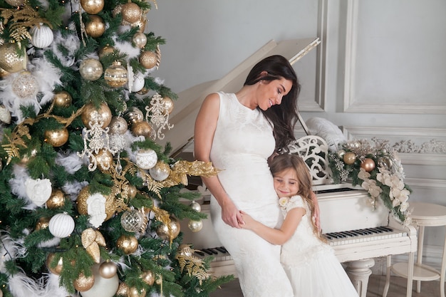 Cheerful mother and her cute daughter girl with a white piano and decorated Christmas tree with presents having fun and hugging in classic luxury New Year interior.