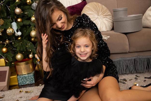 Cheerful mother and her daughter sitting near Christmas tree at home Merry Christmas and Happy Holidays