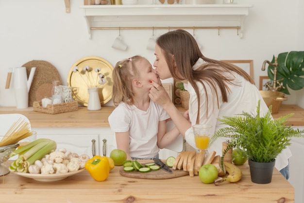 Cheerful mother and little daughter are preparing salad together in the kitchen and having fun. The girl kisses her mother at home.