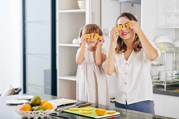 Cheerful mother and little daughter posing with orange slices at kitchen counter