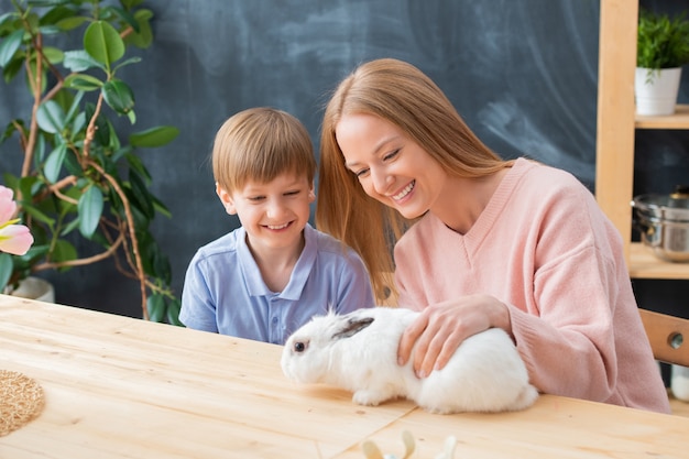 木製のテーブルに座って白いウサギと遊ぶ陽気な母と息子