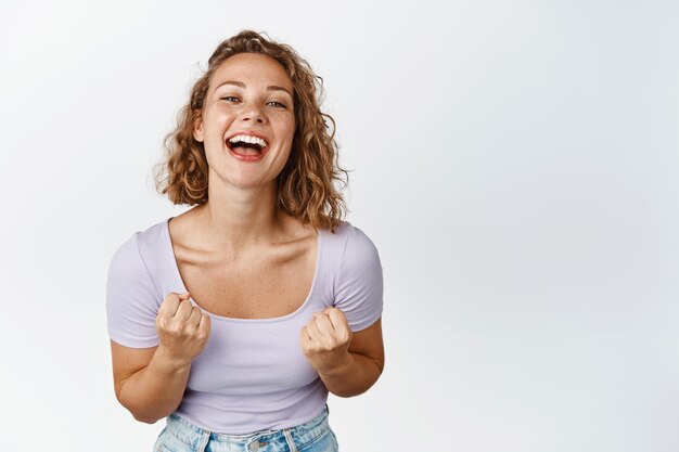 Cheerful and motivated blond girl laughing, smiling and looking happy, clenching fists encouraged, standing over white background