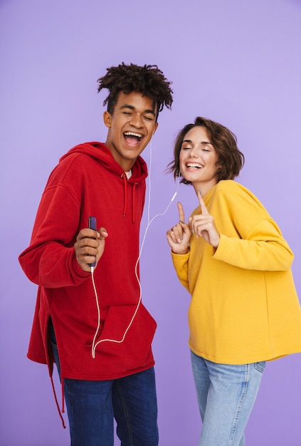 Cheerful multhiethnic teenage couple standing together isolated, listening to music with earphones, holding mobile phone