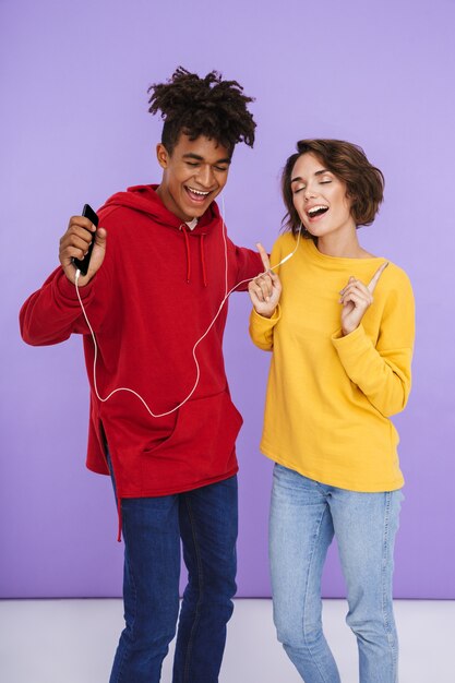 Cheerful multhiethnic teenage couple standing together isolated, listening to music with earphones, holding mobile phone