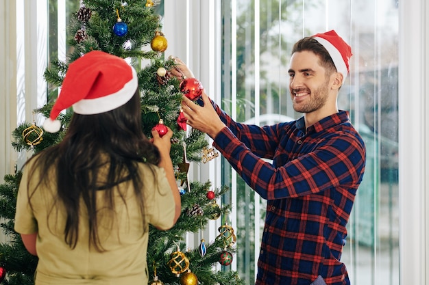 Cheerful multi-ethnic business people decorating Christmas tree in office with colorful bauble
