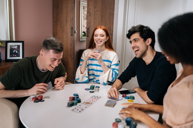 Cheerful multiethnic friends having home fun party on weekends playing poker game sitting at table in living room Diverse laughing men and women satisfied with spending leisure together playing poker