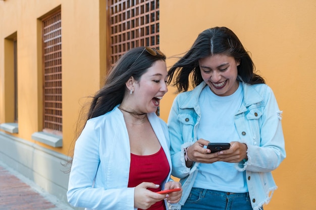 Cheerful multiracial friends in casual clothes laughing and looking at their cell phones as they walk together down a city street on a sunny day.