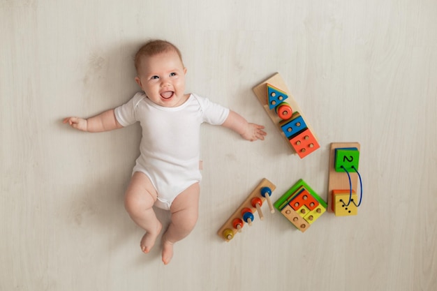 白いボディースーツを着た元気な新生児が床に仰向けになって、知育おもちゃで遊んでいます。子供のための製品。幸せな子供時代と母性の概念。育児。テキスト用のスペース