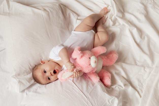 Cheerful newborn baby in a white bodysuit lies on a white bed with a plush pink toy rabbit. products for children, toy. concept of a happy childhood and motherhood. child care. space for text.