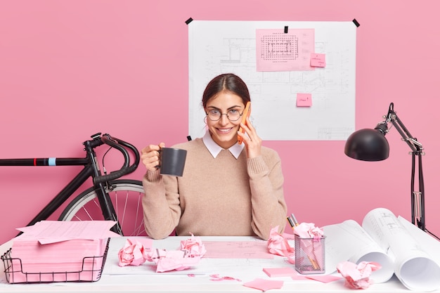 Cheerful office worker works on designing of new office poses in coworking space drinks coffee has telephone conversation 