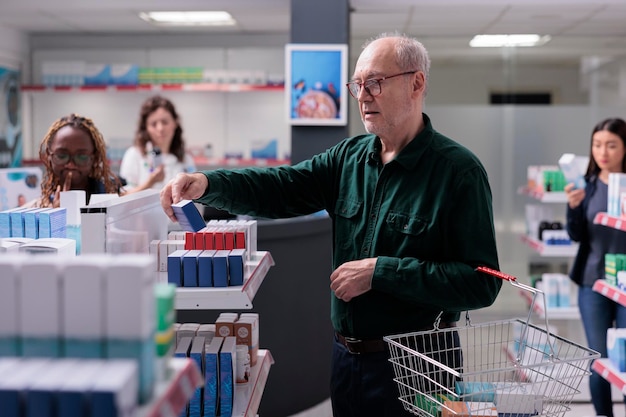 Cheerful old customer looking at pills package, reading
prescription during drugs shopping in pharmacy. elderly man client
choosing to buy supplements, vitamin, health care products