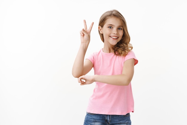 Cheerful optimistic cute blond girl cannot wait play with friends, show peace victory sign, feeling joyful and happy, smiling toothy positive expression