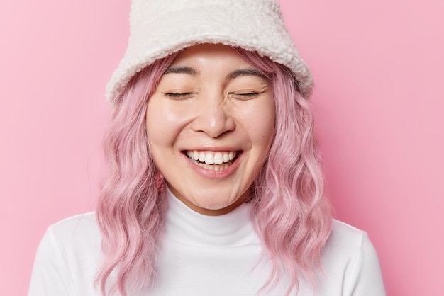 写真 バラ色の髪の陽気な大喜びのアジアの女性は心から笑う白い歯が目を閉じていることを示していますピンクの背景の上に帽子とポロネックを身に着けています人々の感情と感情の概念
