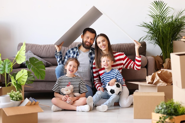 Cheerful parents and kids holding house roof over head and smiling for camera while sitting near couch and belongings in new flat