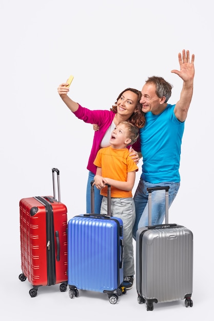 Cheerful parents and son smiling and posing for selfie while standing behind suitcases before trip against white background