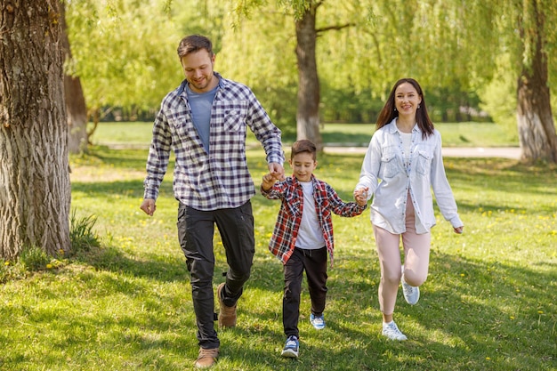 Cheerful parents with small son walking hand in hand in green summer park Happy family