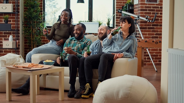 陽気な人々がビールのボトルをチリンと鳴らし、テレビチャンネル番組で映画を見て、ハウスパーティーで一緒に楽しんでいます。テレビで映画を見ながらお酒を飲む友人。