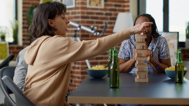 Cheerful people having fun with society games at home, playing with wooden construction structure cubes and square building pieces. Modern group of friends enjoying competition.