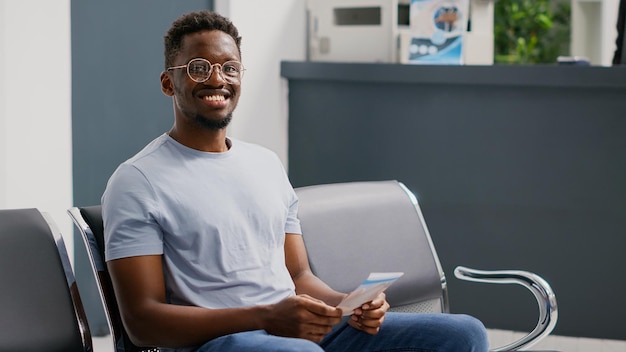 Cheerful person waiting in facility hallway to meet with physician, sitting in waiting area before medical consultation. Happy male patient at reception area having checkup appointment.