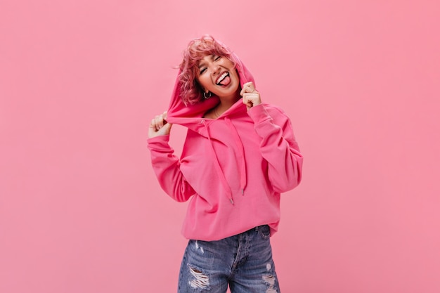 Cheerful pink-haired woman in fuchsia hoodie and denim shorts demonstrates tongue and makes funny face on isolated wall 