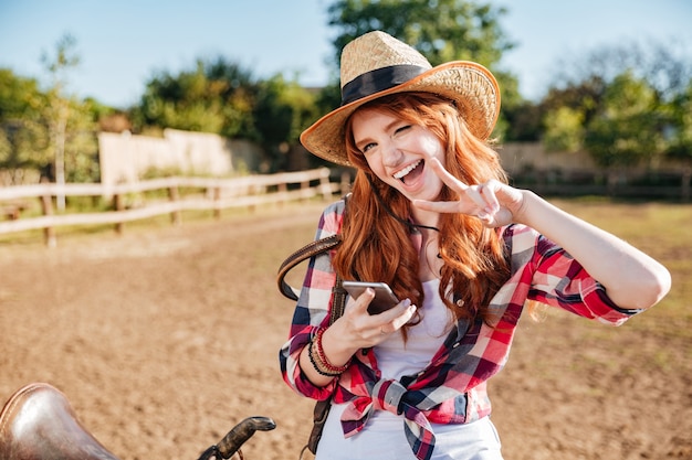 Cheerful playful young woman cowgirl in hat using cell phone and showing peace sign