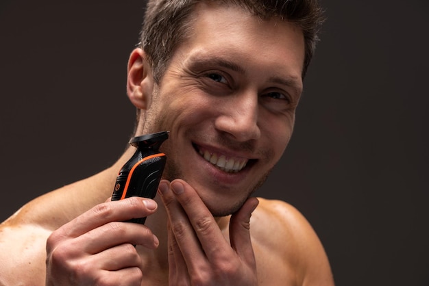 Cheerful portrait of man trimming his beard. Young caucasian man with modern electric beard trimmer. Stock photo