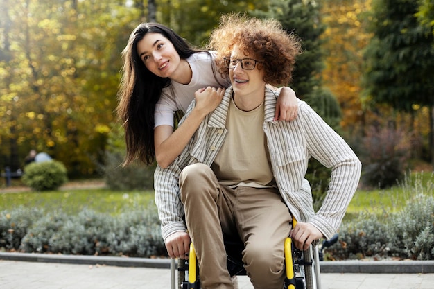 Foto allegra ragazza bruna positiva abbraccia la sua amica un giovane sorridente si siede su una sedia a rotelle per strada