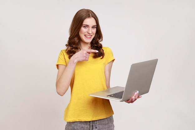 Cheerful positive teenager girl in yellow casual style T-shirt pointing finger at display of laptop, blogger with portable computer. Indoor studio shot isolated on gray background.