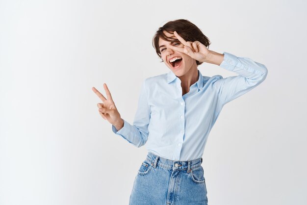 Donna positiva allegra divertendosi sorridendo e ridendo mostrando segni di pace sull'occhio in piedi su sfondo bianco