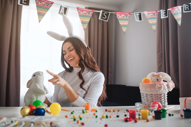 Cheerful positive young woman prepare for Easter alone. She sit at table in room and play with white rabbit toy. Decoration paint and sweets colorful on table. Woman wear festive ears.